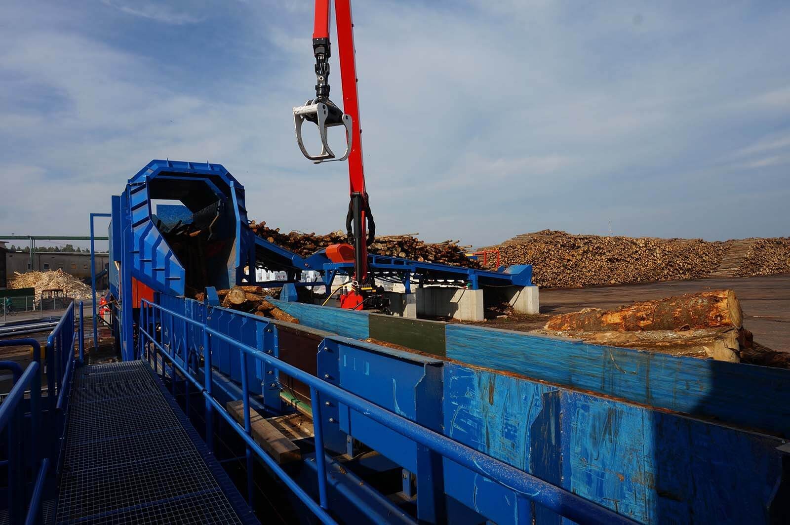 Grue à grappin chargeant des billons dans la goulotte d'une ligne d'écorçage, parc à bois à l'arrière-plan
