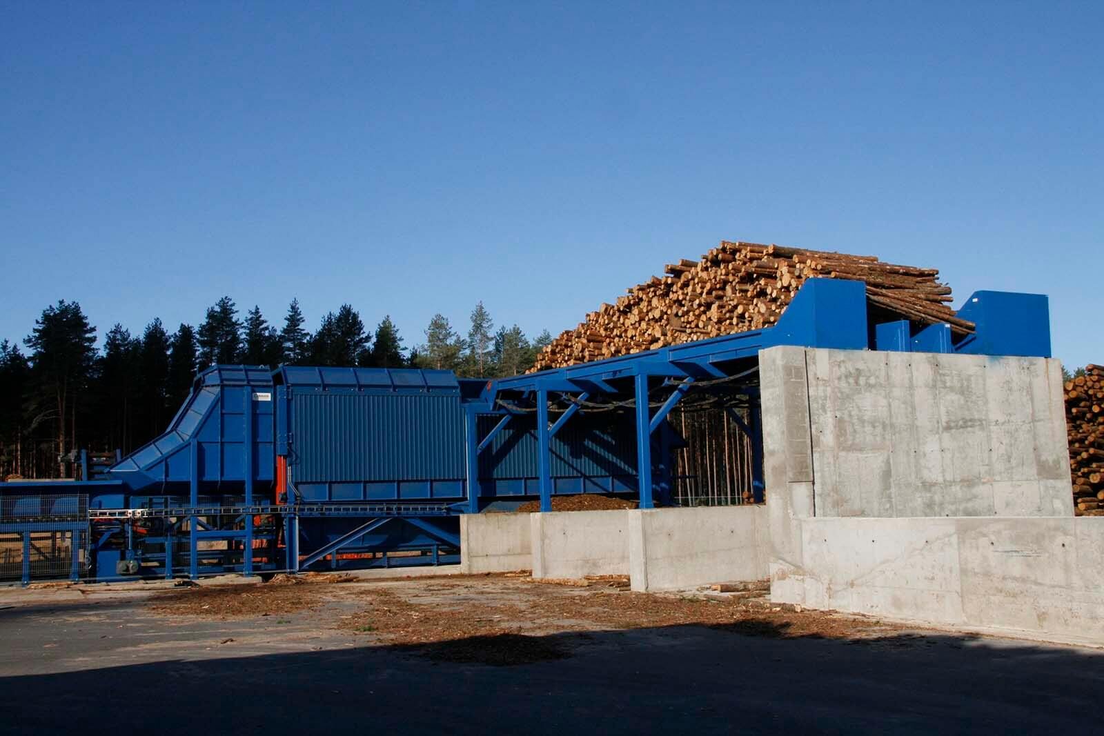 Ligne d'écorçage bleue en extérieur&nbsp;: table d'alimentation chargée de bois ronds sur des murs en béton, pins en fond